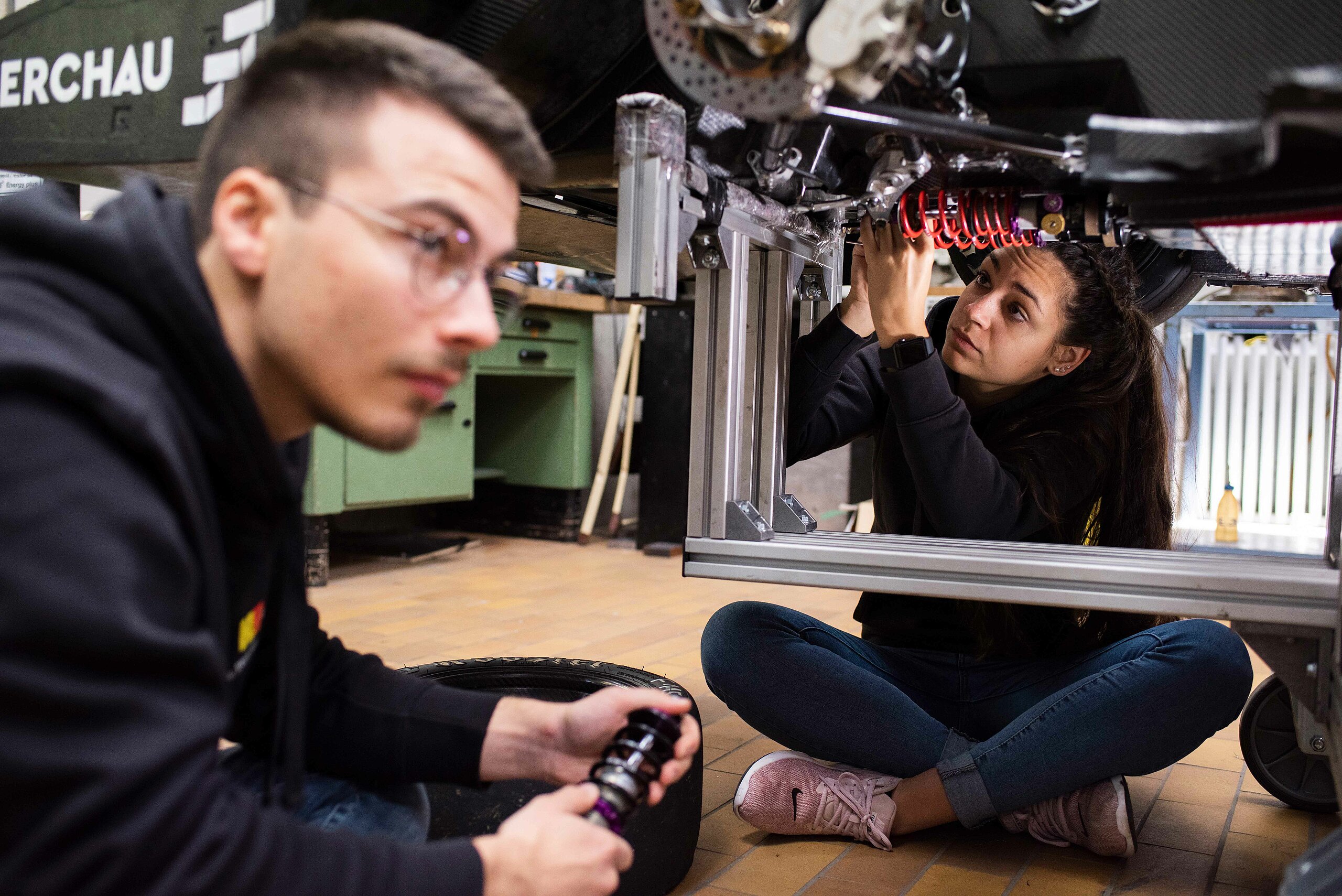 two students sit below a metal frame bearing wires and work on it