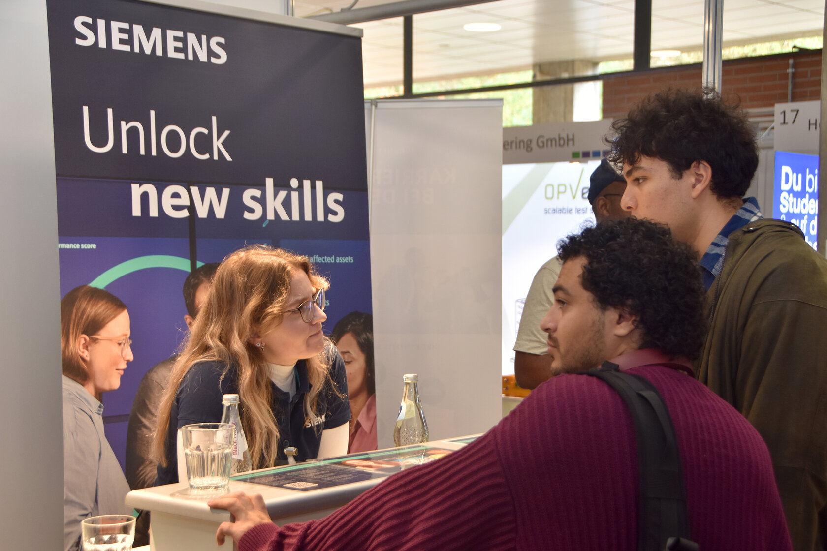 Zwei Studenten sprechen mit einer Siemens-Mitarbeiterin an einer Messetheke. Im Hintergrund steht ein großes Siemens-Banner mit der Aufschrift „Unlock new skills“.