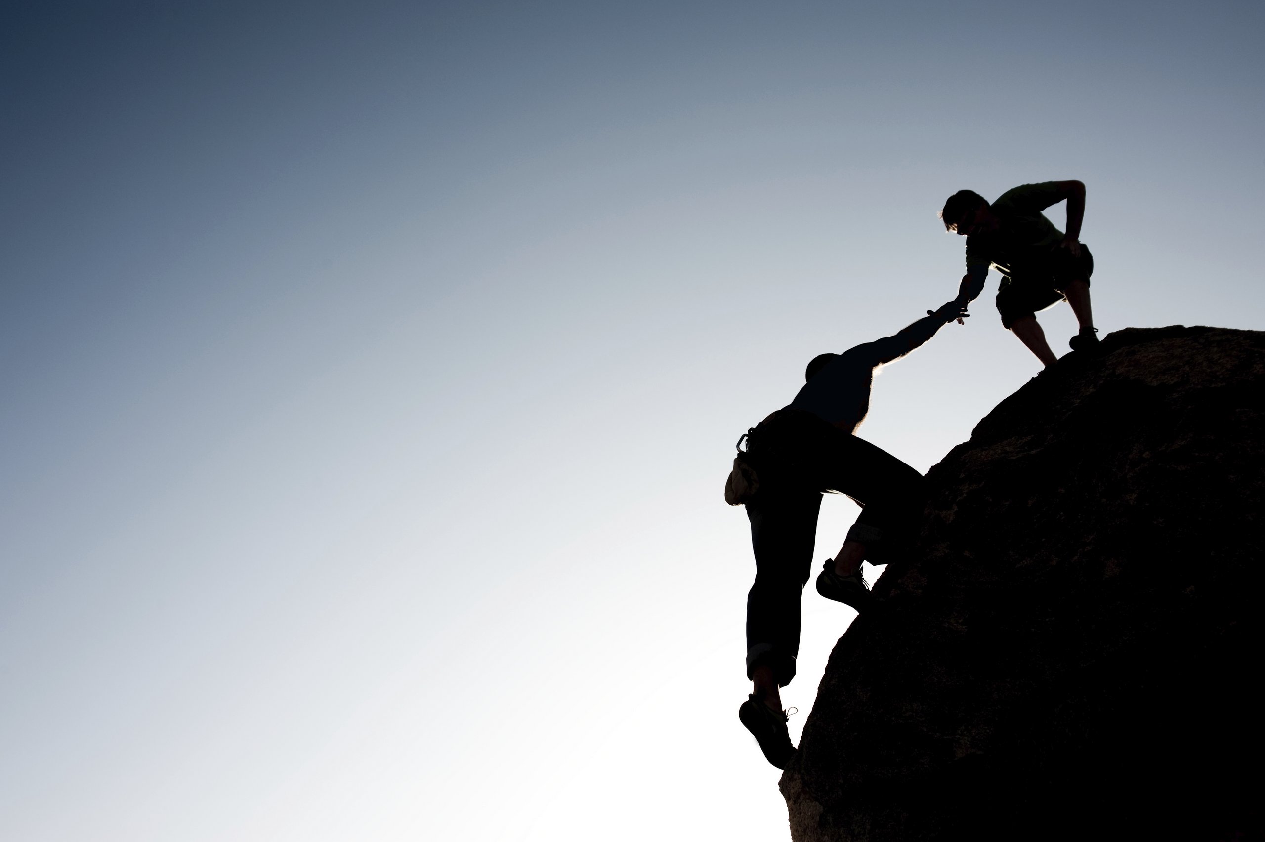 Eine Person steht auf einem Berg, reicht einer zweiten Person die Hand und hilftihr so, auf den Berg zu kommen. 
