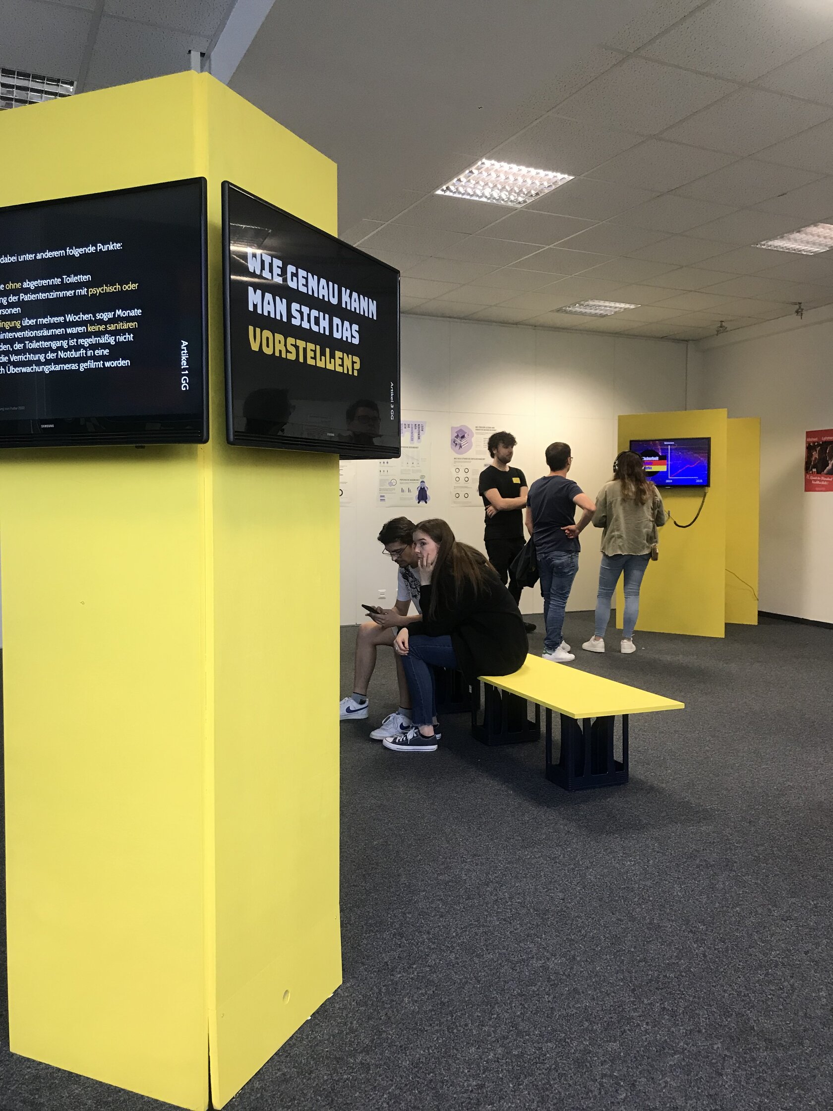 Raum mit verschiedenen Stationen, Säulen mit Bildschirmen und einer Bank: „Grundgesetzte – Theorie vs. Realität“ von Jana Exner, Dalina Hampp, Ian Klemm
