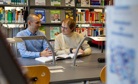 Zwei Studierende sitzen in der Bibliothek und unterhalten sich über ein Buch gebeugt.