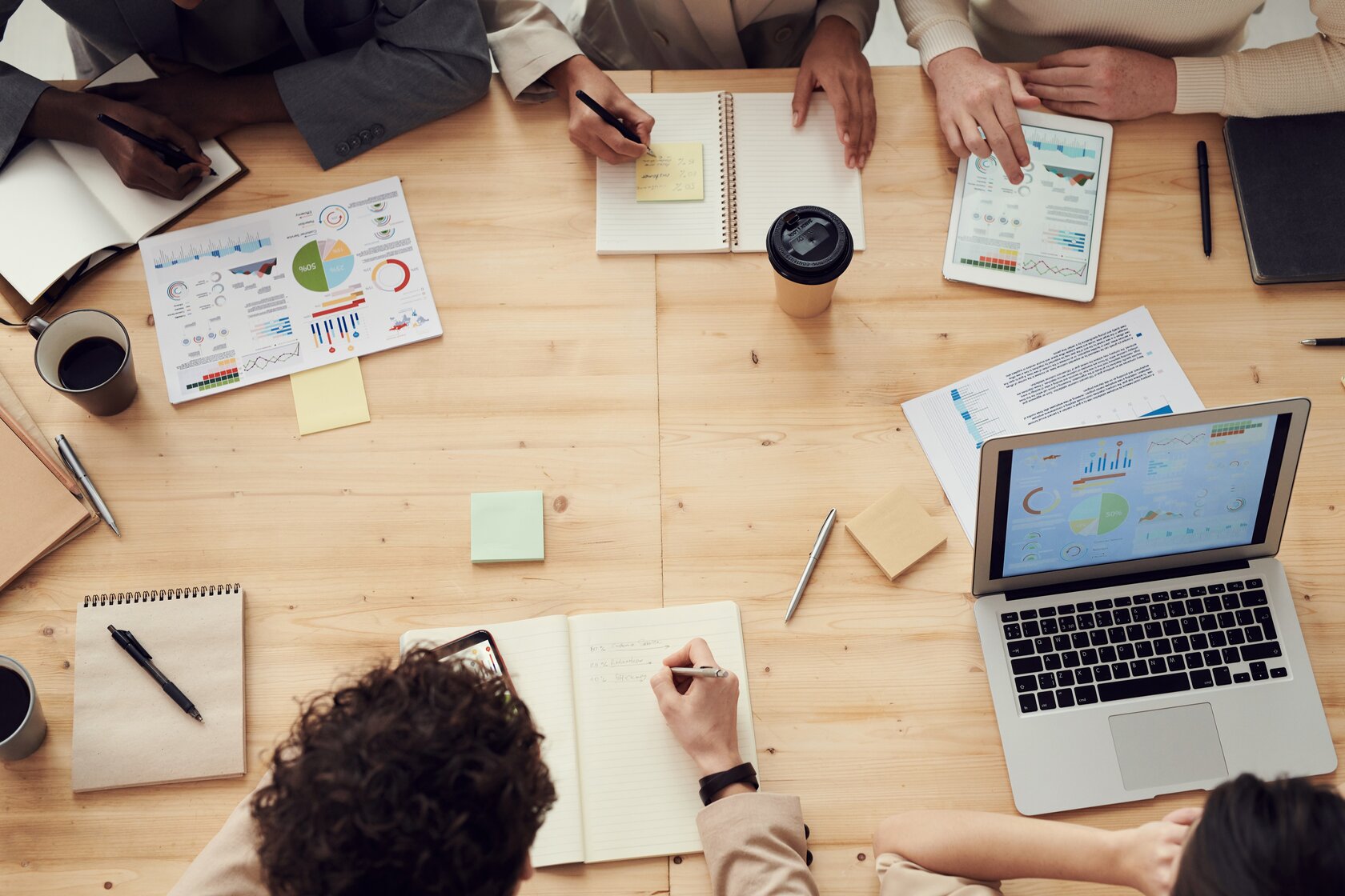 table from above, with people sitting around it, and writing pads, open laptop, and coffee cups strewn across