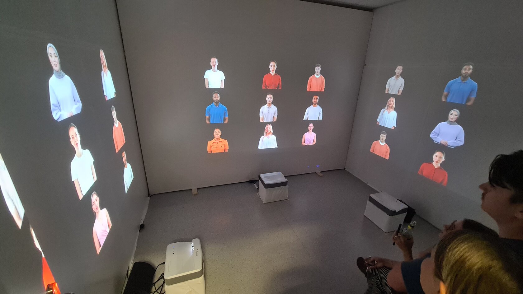 In einem kleinen Raum sind verschiedene Personen an die Wände projiziert worden. „Illusion oder Realität: Wie Deepfakes unser Vertrauen in die Wahrheit erschüttern“ von Lea Karch, Antonia Rademacher, Pauline Fuchs, Senta Hamm, Samira Weigand, Noelle Wilser