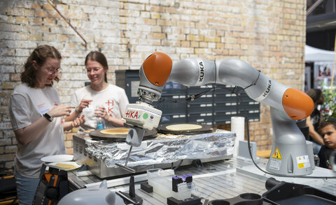 Roboterarm mit der Aufschrift „KUKA“ und „HKA“ bereitet auf einer Crêpe-Platte einen Crêpe zu, im Hintergrund zwei Personen in weißen HKA-T-Shirts vor einer Backsteinwand.