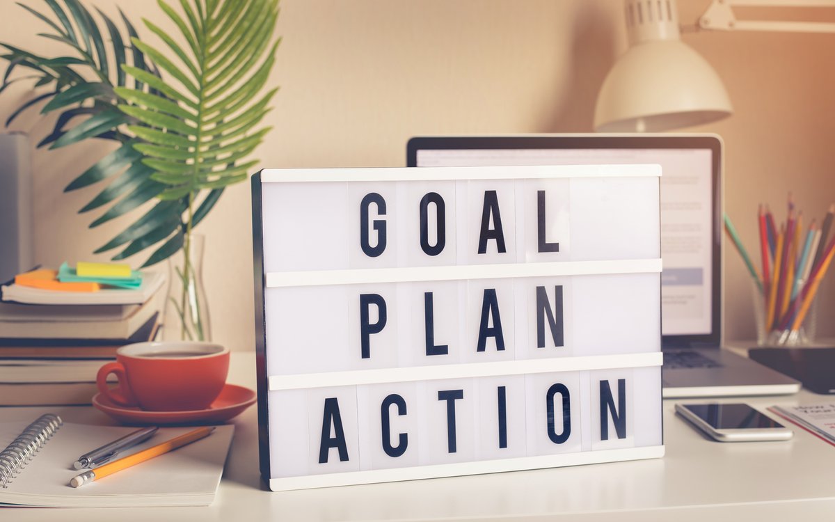 [Translate to English:] Schreibtisch mit einem Schild, auf dem die Worte Goal, Plan, Action zu lesen sind.