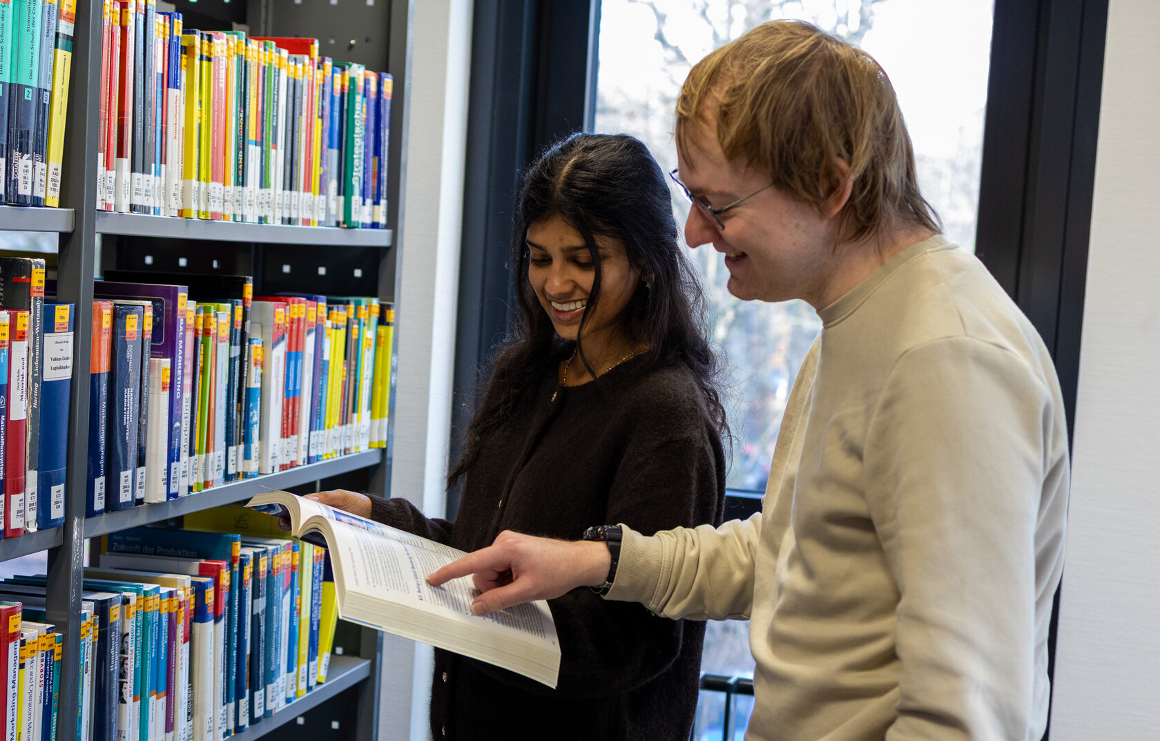 Zwei Studierende vor einem Bücherregal halten ein Buch und unterhalten sich.