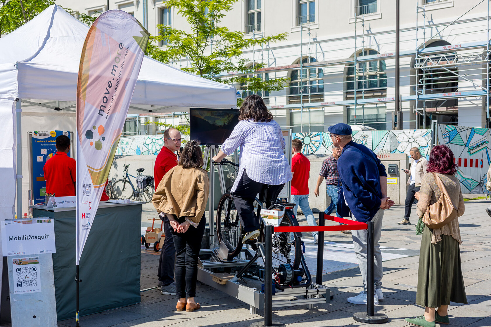 Eine Frau fährt auf einem stationären E-Bike-Simulator mit Bildschirm, umgeben von mehreren Zuschauenden vor dem move.mORe-Stand.