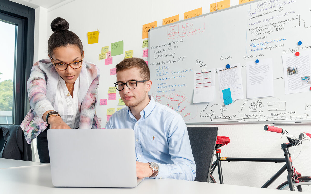 two students working at laptop, behind them a whiteboard covered in sticky notes and writing