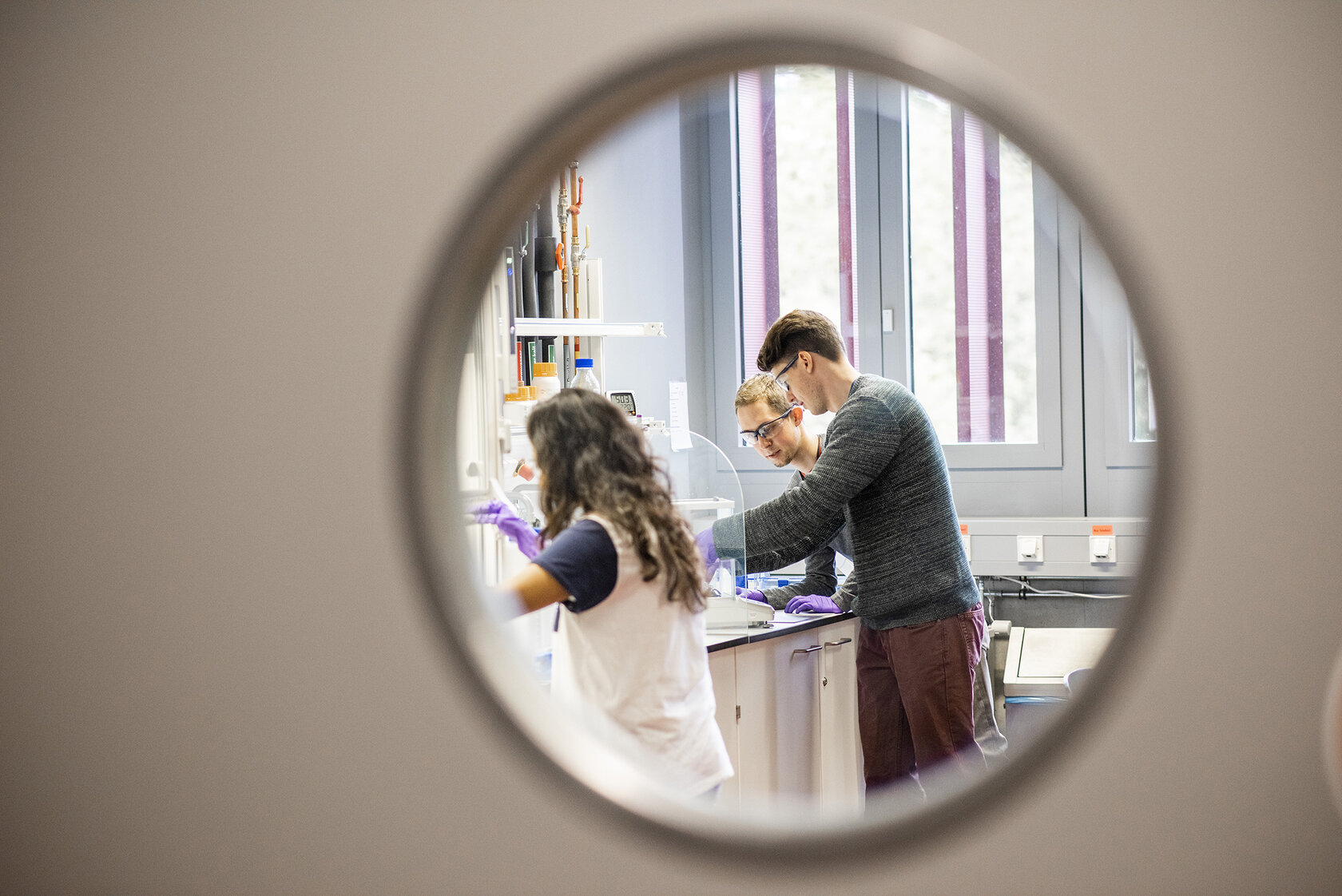 three young people making tests at a workstation, seen through a round window