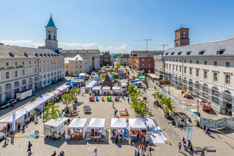 Luftaufnahme des Karlsruher Marktplatzes mit zahlreichen weißen Pavillons, bunten KLIMA-Großbuchstaben und der Stadtpyramide bei blauem Himmel.