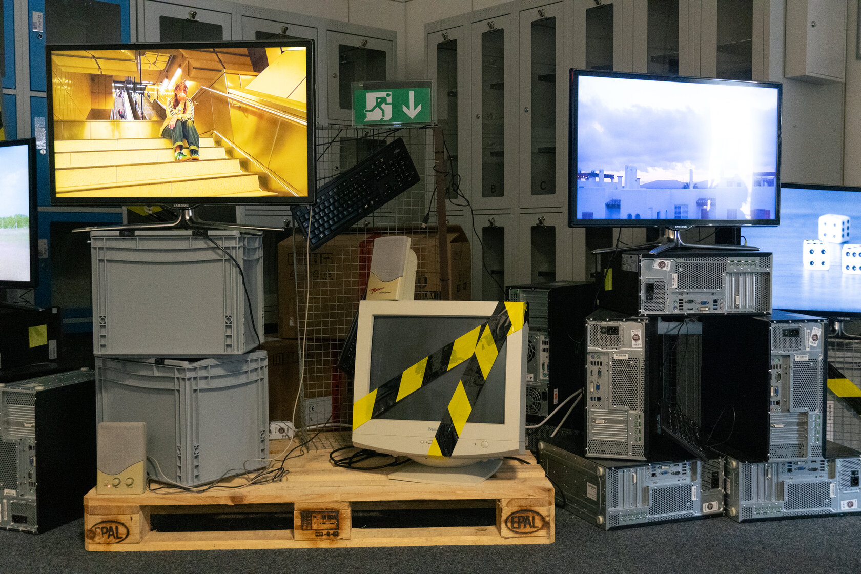 Ausstellungsstück „Chronos“ von Norea Börnsen, Antonia Linke, Amelie Vatterott und Liv Walter: mehrere Monitore und Computer, teils mit ablaufenden Videos, arrangiert auf einer Europalette mit Euroboxen und Absperrband