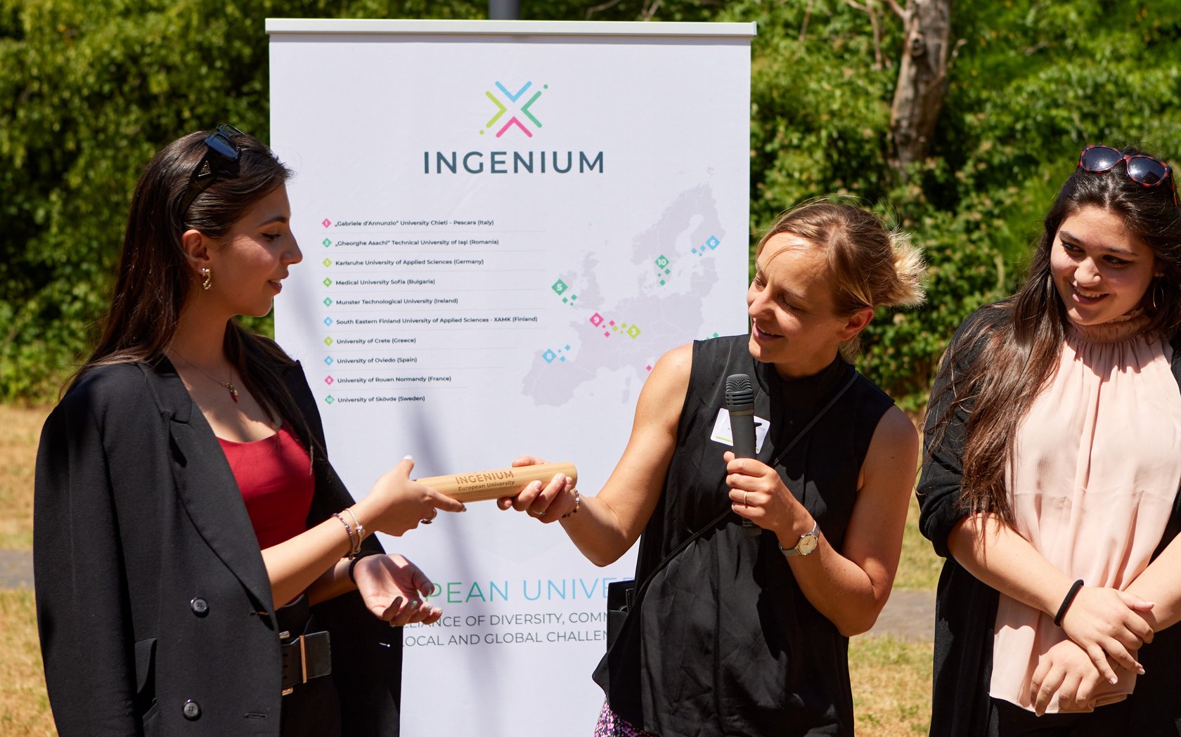 Ms Rüppell-Wee standing in front of INGENIUM rollup with two students, handing over a baton