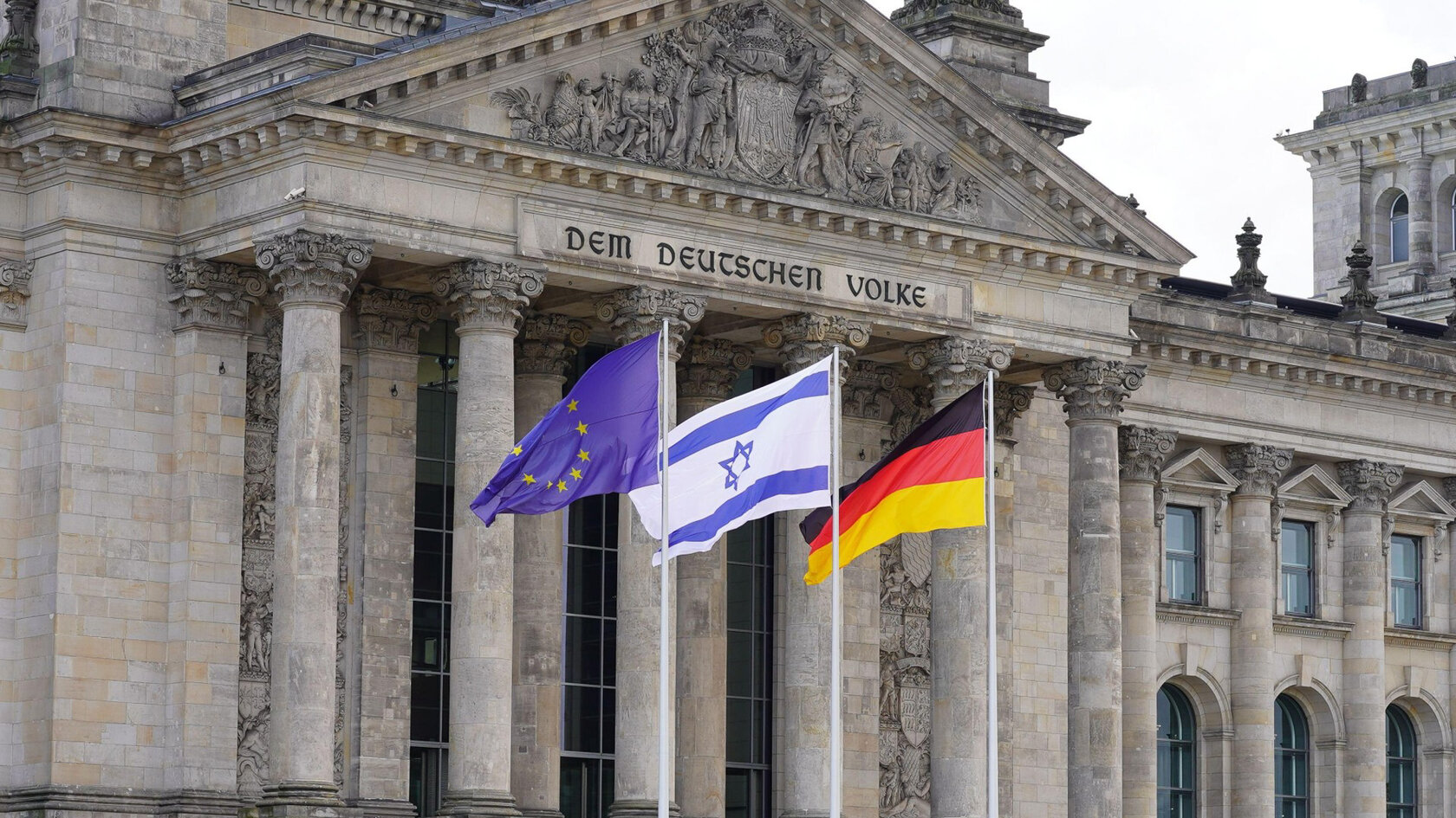 Blick auf den Eingangsbereich des Reichstagsgebäudes in Berlin. Vor dem Säulenportal wehen drei Flaggen: die blaue EU-Flagge mit goldenen Sternen, die weiß-blaue israelische Flagge mit dem Davidstern sowie die schwarz-rot-goldene Deutschlandfahne.