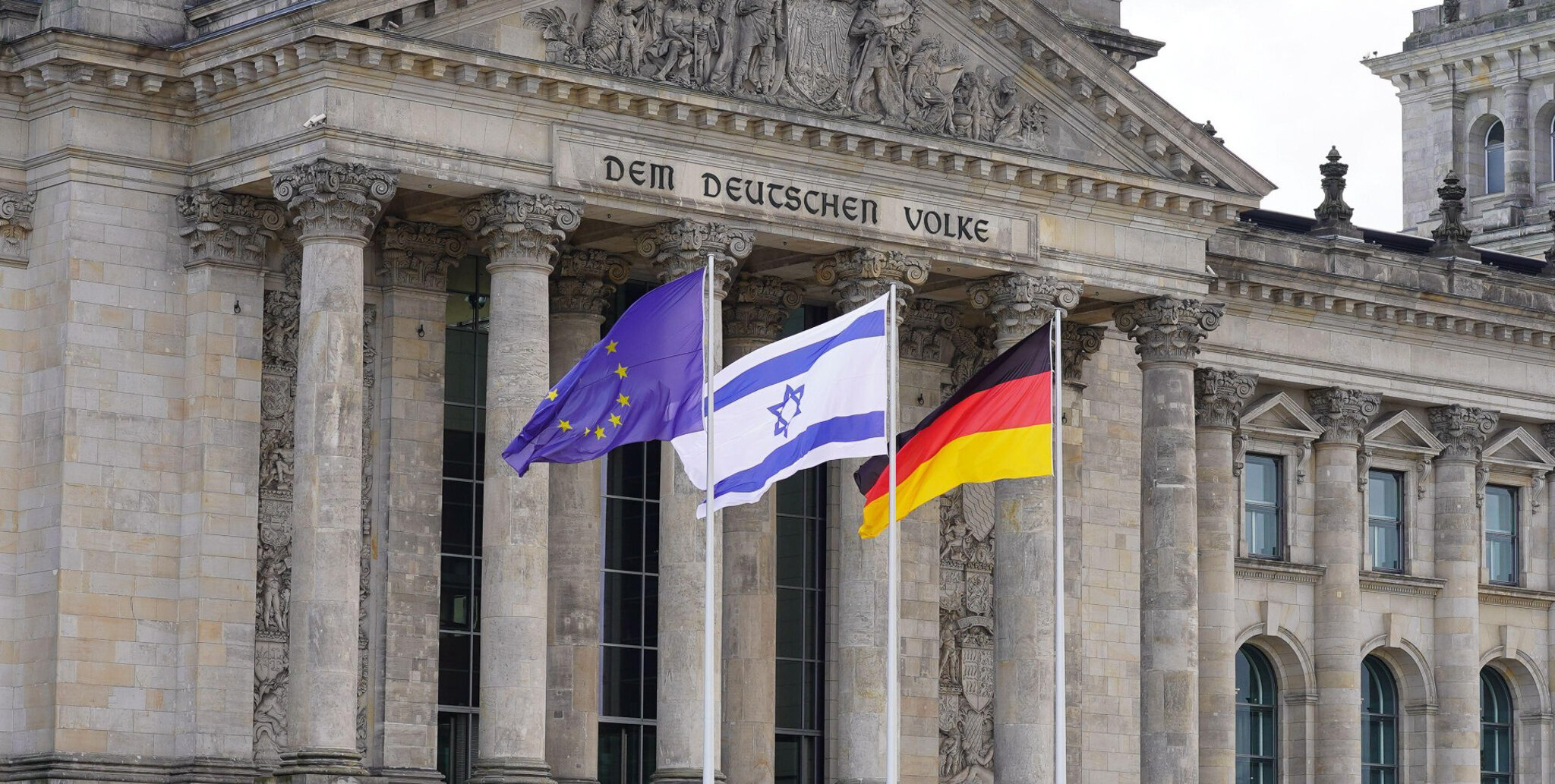 Blick auf den Eingangsbereich des Reichstagsgebäudes in Berlin. Vor dem Säulenportal wehen drei Flaggen: die blaue EU-Flagge mit goldenen Sternen, die weiß-blaue israelische Flagge mit dem Davidstern sowie die schwarz-rot-goldene Deutschlandfahne.