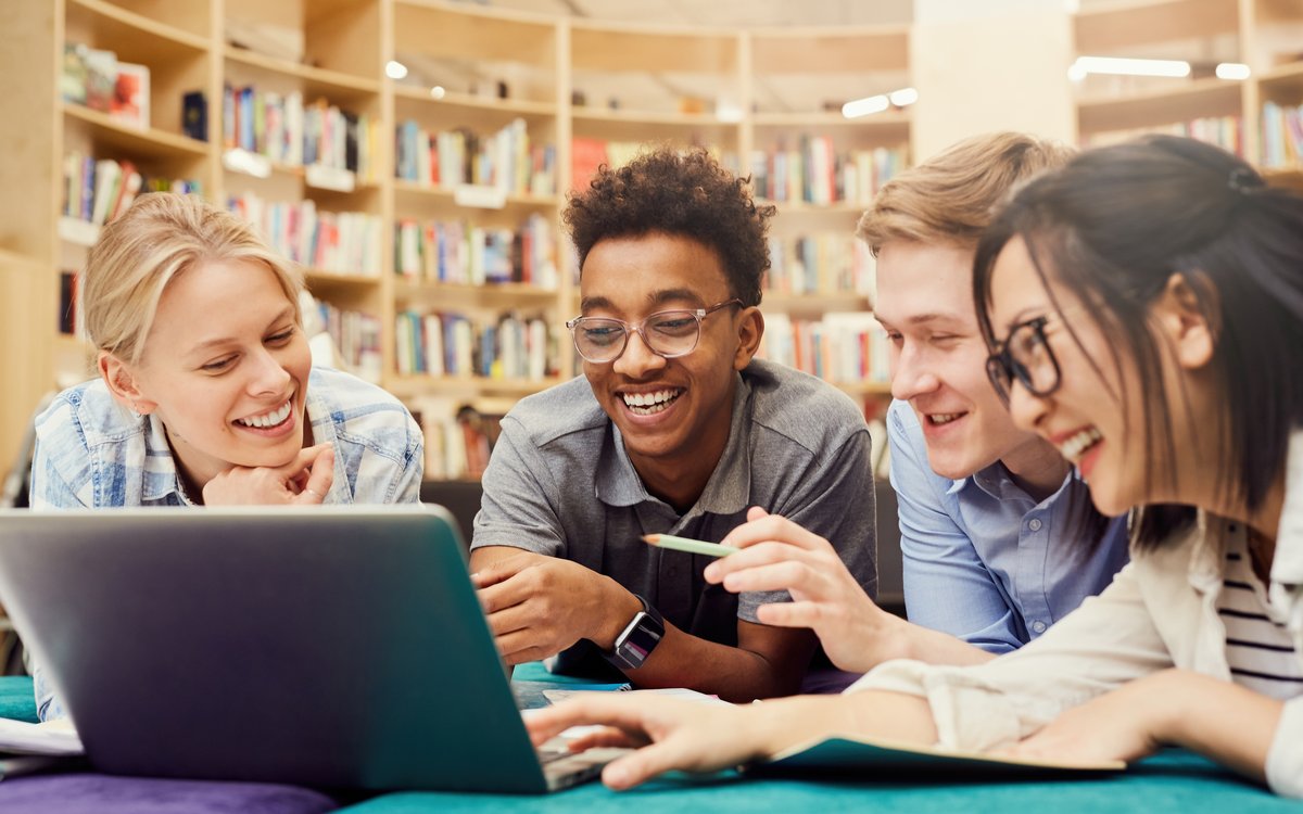 Multiethnische Studierende liegen mit einem Laptop auf dem Boden einer Bibliothek und lächeln.