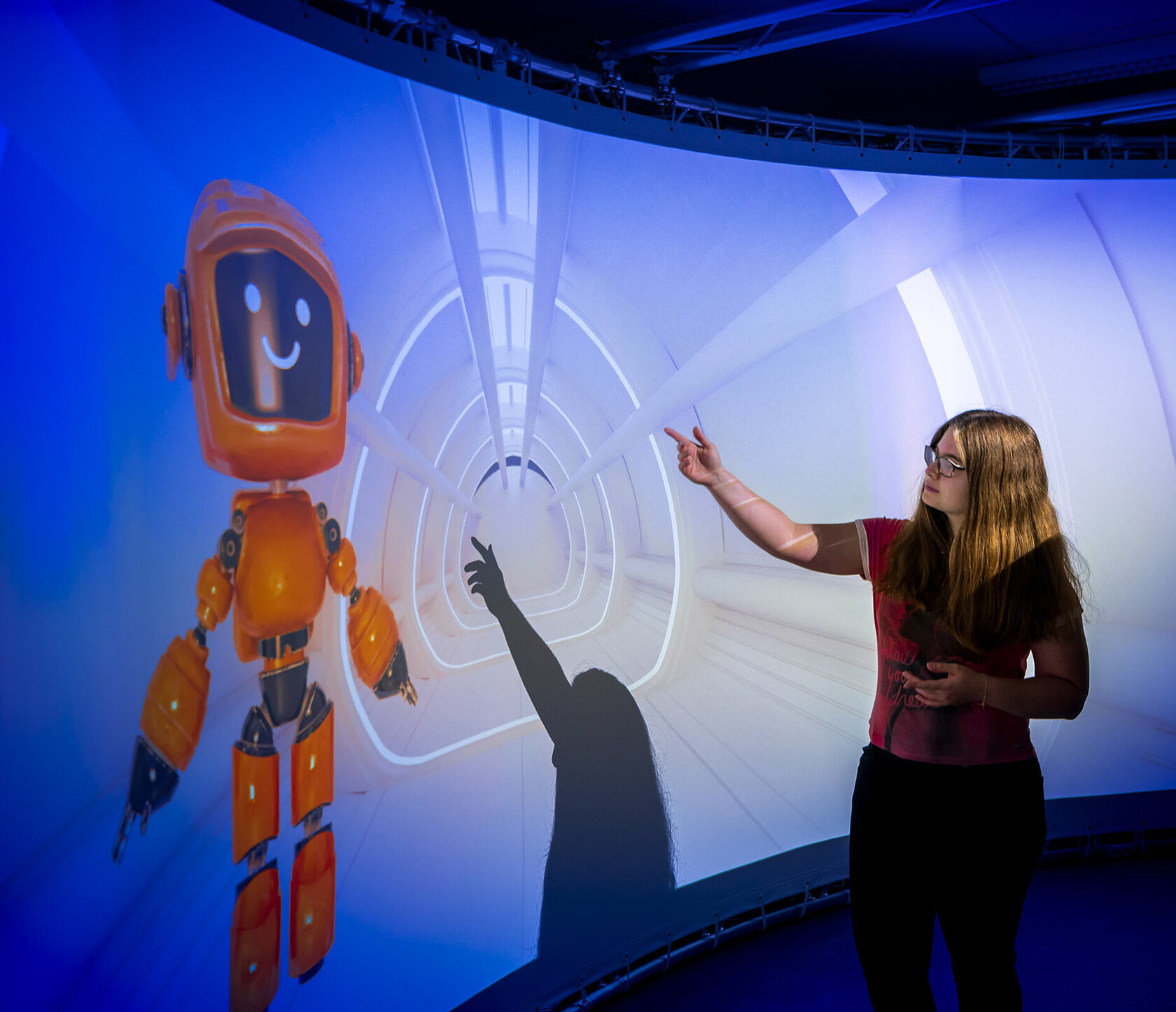 The image shows a young woman interacting with a friendly orange robot in an immersive projection environment. The robot, an "embodied conversational agent", floats in a futuristic-looking tunnel setting and has a black display face with a smiling expression. The woman points at the digital assistant with an outstretched arm, while her shadow also falls on the projection and appears to touch the robot.