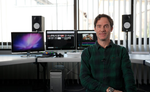 Prof. Sebastian Krügler sitzt vor einem Schreibtisch mit vielen Monitoren und Lautsprechenern. Auf den Bildschirmen ist ein Videoprojekt zu sehen, dass sich gerade im Schnitt befindet.
