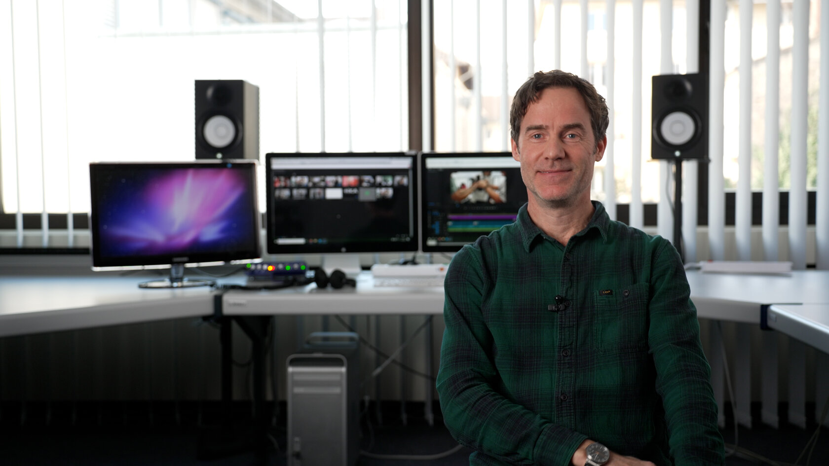 Prof. Sebastian Krügler sitzt vor einem Schreibtisch mit vielen Monitoren und Lautsprechenern. Auf den Bildschirmen ist ein Videoprojekt zu sehen, dass sich gerade im Schnitt befindet.