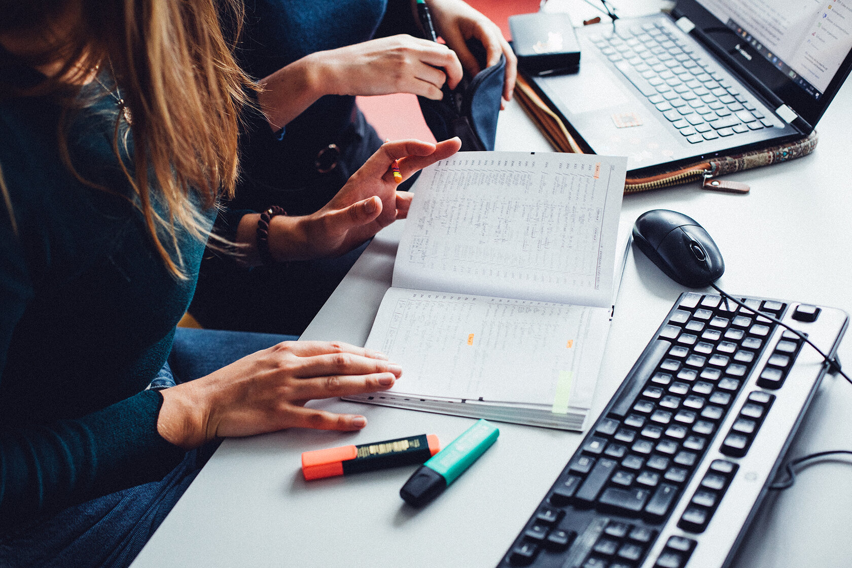 Blick auf einen Schreibtisch mit Laptop, Maus, Tastatur und Stiften und auf die Hände einer Studentin, die in einem Schreibblock blättert