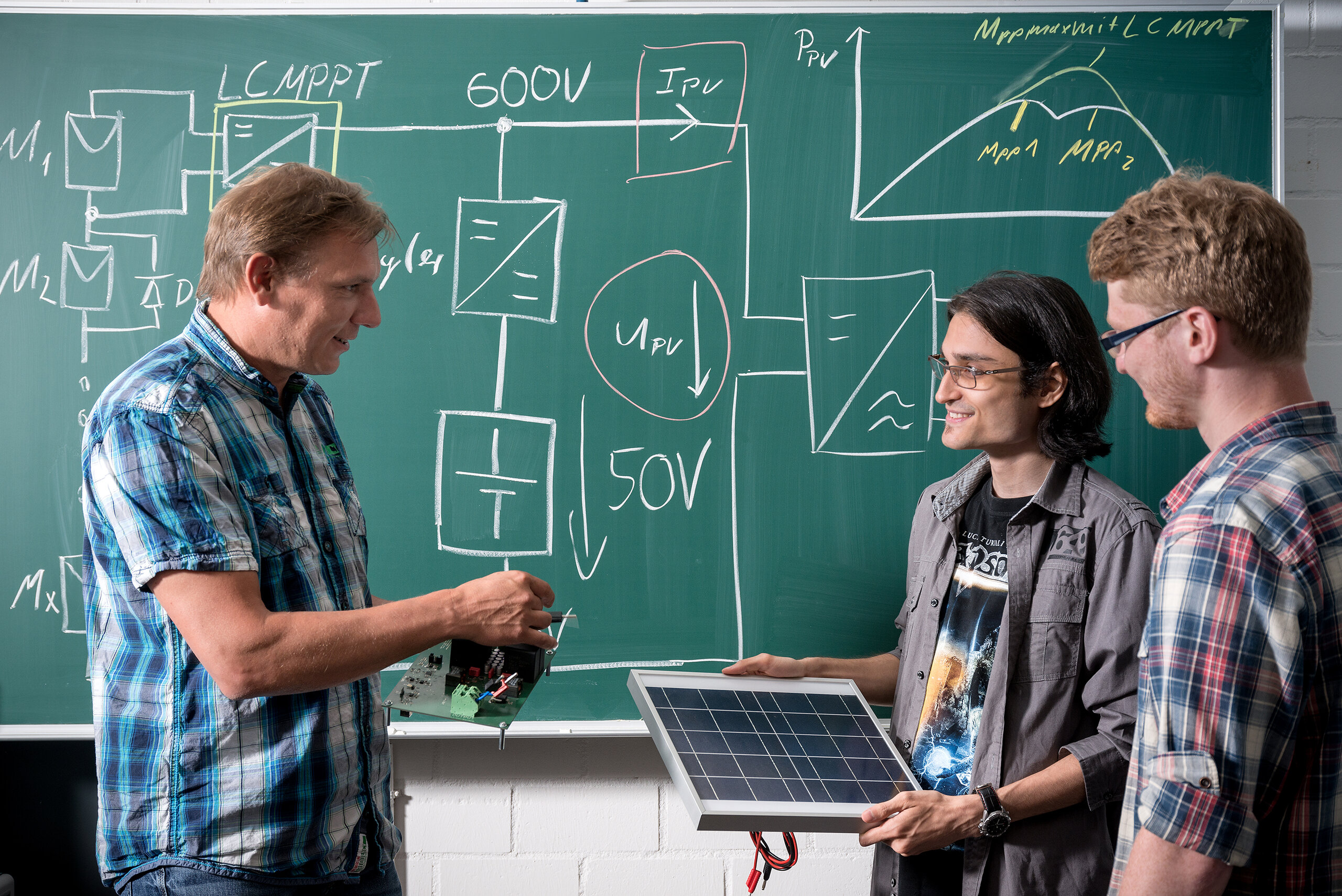 lecturer talking with two students standing in front of blackboard covered in technical diagrams