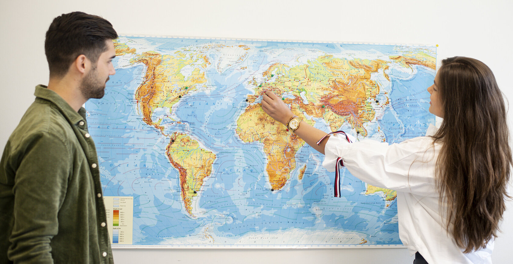 two students standing in front of a world map on the wall