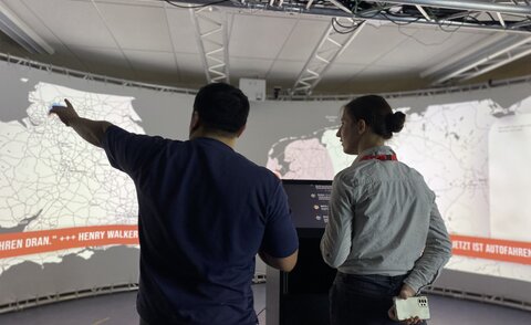Two people stand in front of a large curved projection screen in the iXperience Lab and watch an interactive visualization on a 180-degree screen. One person points to a map while explaining the application.