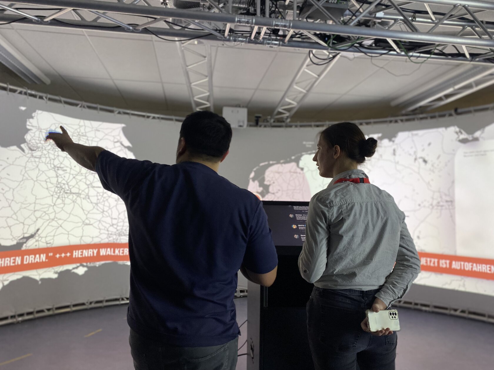 Two people stand in front of a large curved projection screen in the iXperience Lab and watch an interactive visualization on a 180-degree screen. One person points to a map while explaining the application.