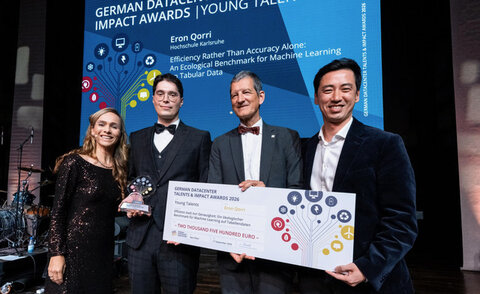 Eron Qorri is standing on stage with three other people, holding his award and a symbolic check. In the background, his name can be seen on the event screen.