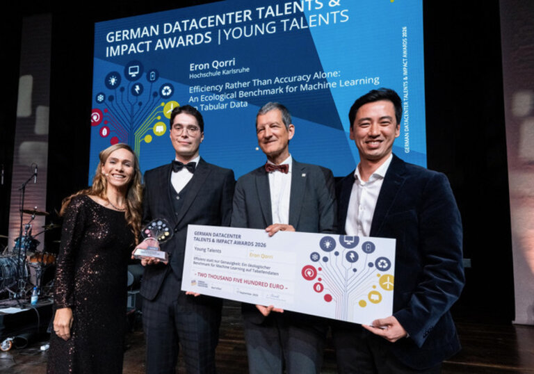 Eron Qorri is standing on stage with three other people, holding his award and a symbolic check. In the background, his name can be seen on the event screen.