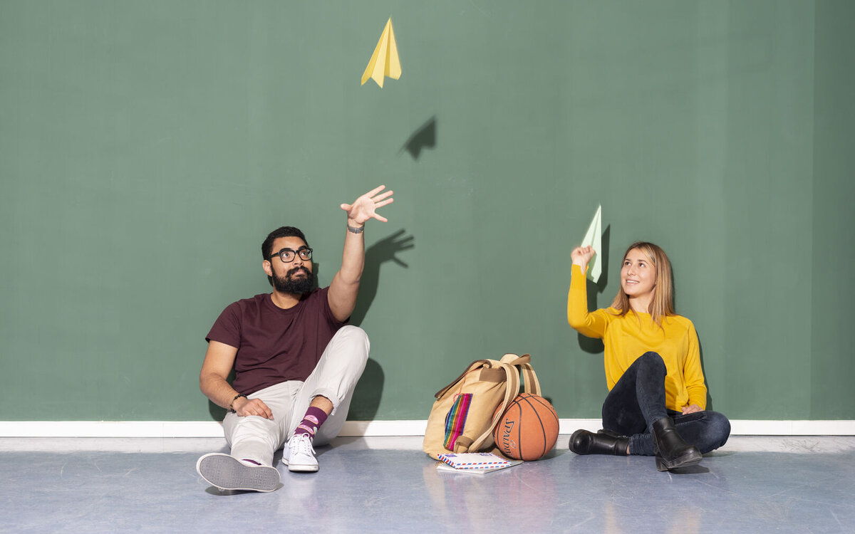 two young people sitting against a wall and letting fly paper airplanes