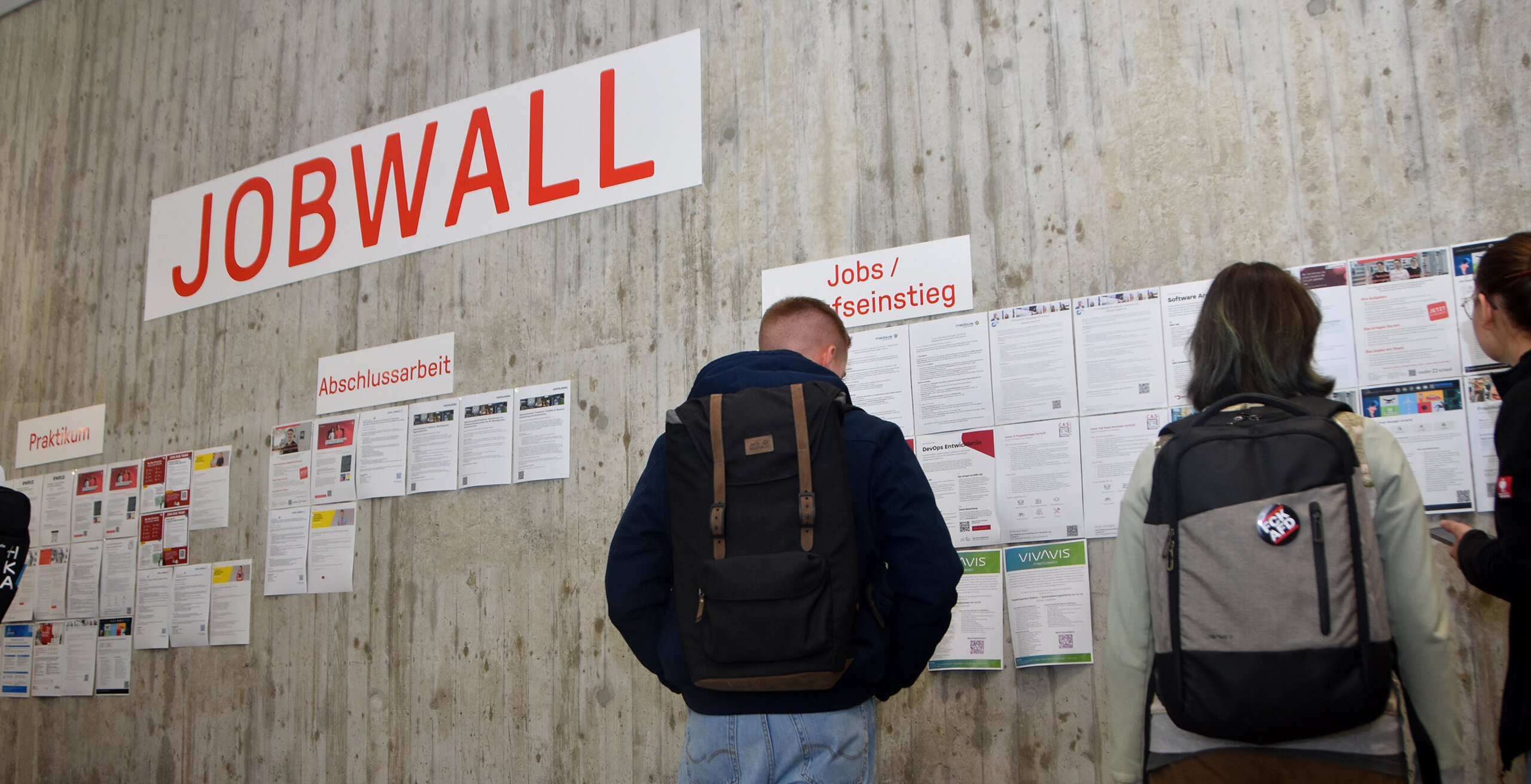 Studierende betrachten zahlreiche Stellenausschreibungen an einer Betonwand. Darüber hängen Schilder mit den Aufschriften „Jobwall“, „Praktikum“, „Abschlussarbeit“ und „Jobs / Berufseinstieg“.