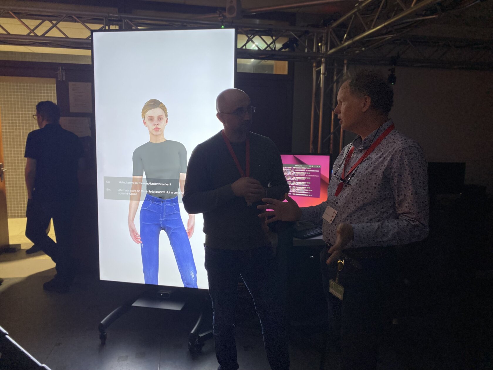 Two visitors stand in front of a large screen with a virtual avatar. The avatar is part of an embodied conversational agent that is being developed in the iXperience Lab and can interact with people.