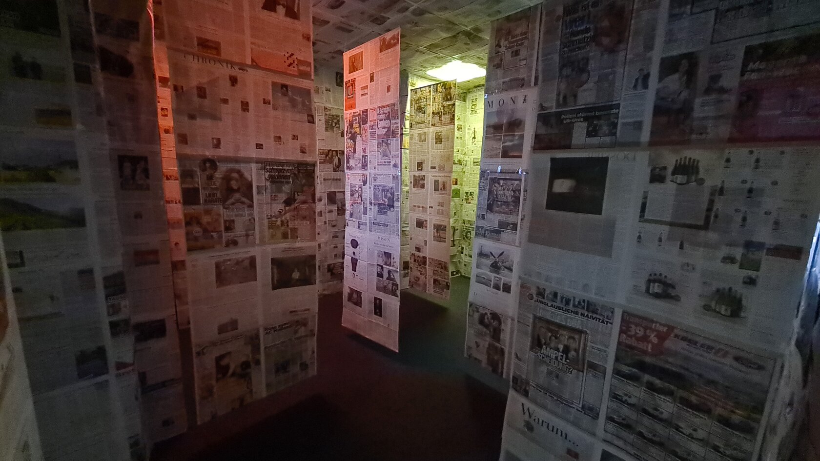 „Zwischen Fakten und Fake News“ von Nina Baum, Gianluca Giuditta, Lionel Kelly, David Krauß, Jana Weigelt zeigt einen Raum mit Zeitungen an den Wänden und weiteren Zeitungen, die wie ein Vorhang von der Decke hängen.