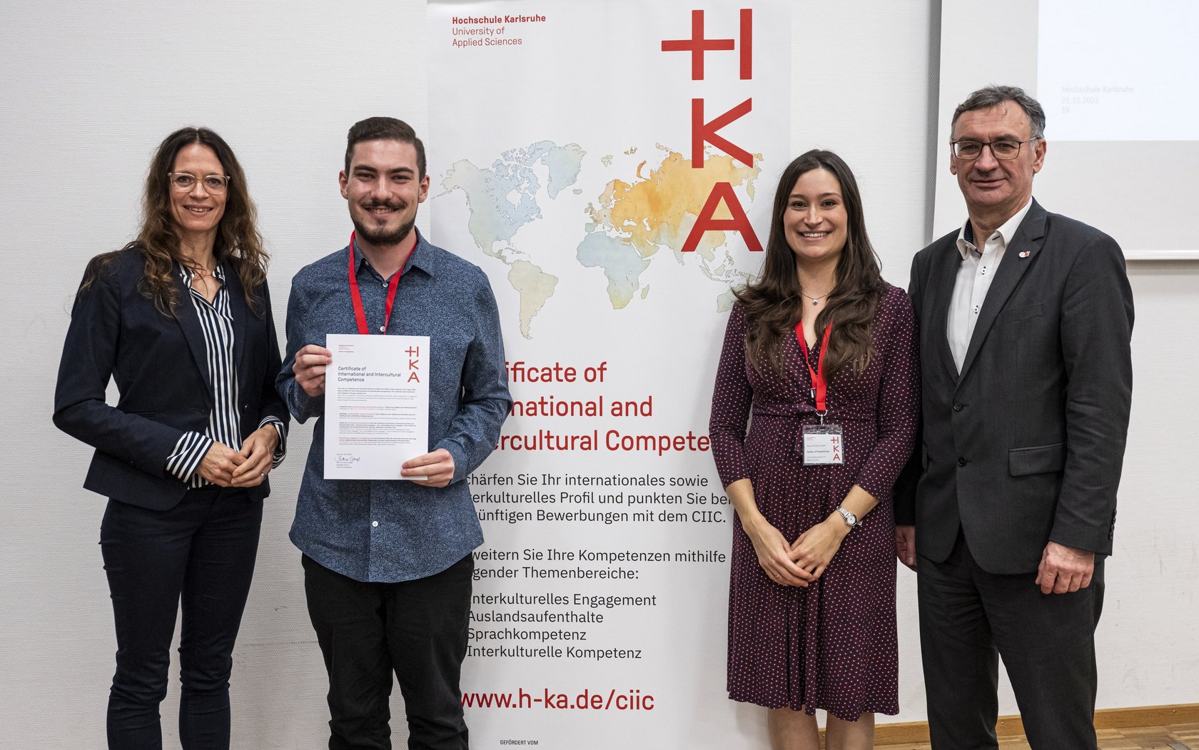 student standing in front of CIIC rollup, presenting his certificate, and surrounded by HKA representatives
