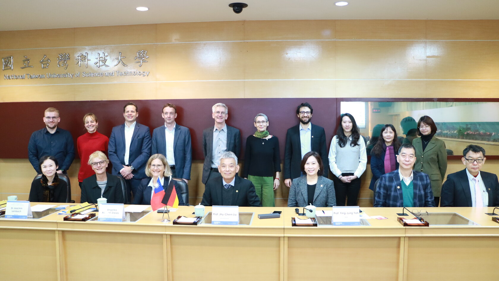 row of people sitting at a table in a conference hall, second row standing behind them