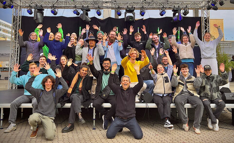Die 26 Studierenden der Hochschule Karlsruhe und ihre Betreuer stehen auf einer Bühne der Local Conference of Youth 2024 in Berlin und reißen in bester Stimmung die Arme nach oben