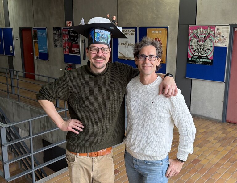 Daniel Hepperle und Matthias W&ouml;lfel stehen nebeneinander und haben sich den Arm gegenseitig &uuml;ber die Schulter gelegt. Beide lachen in die Kamera. Daniel Hepperle steht links und tr&auml;gt einen Doktorhut aus Pappe, auf dem unter anderem das 3D-Modell einer VR-Brille und ein 3D-Modell seines VR-Avatars zu sehen sind, au&szlig;erdem ist der Hut mit Bildern vom IIIX-Team beklebt. 