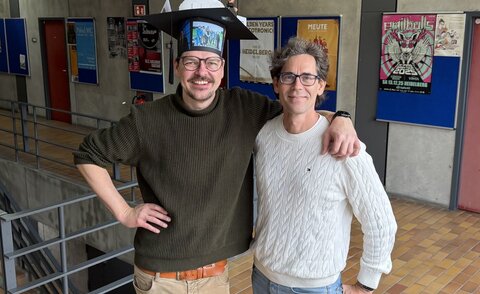 Daniel Hepperle and Matthias W&ouml;lfel are standing next to each other with their arms over each other's shoulders. Both are laughing into the camera. Daniel Hepperle is standing on the left and is wearing a cardboard doctor's hat on which, among other things, a 3D model of VR glasses and a 3D model of his VR avatar can be seen; the hat is also covered with pictures of the IIIX team. 