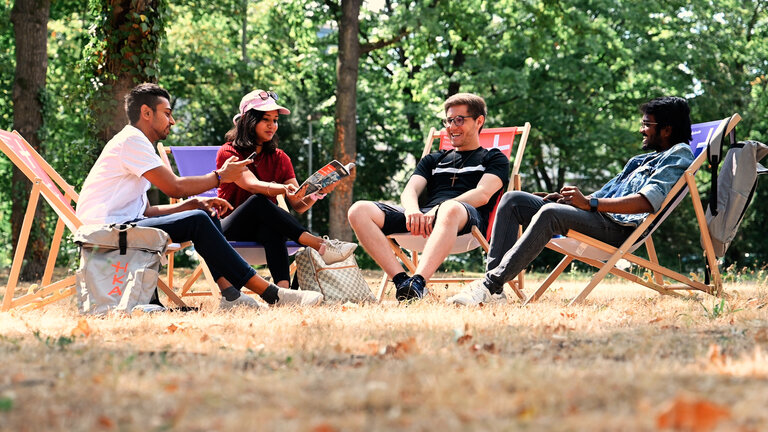 Vier internationale Studierende sitzen auf Liegest&uuml;hlen im Park und unterhalten sich. 