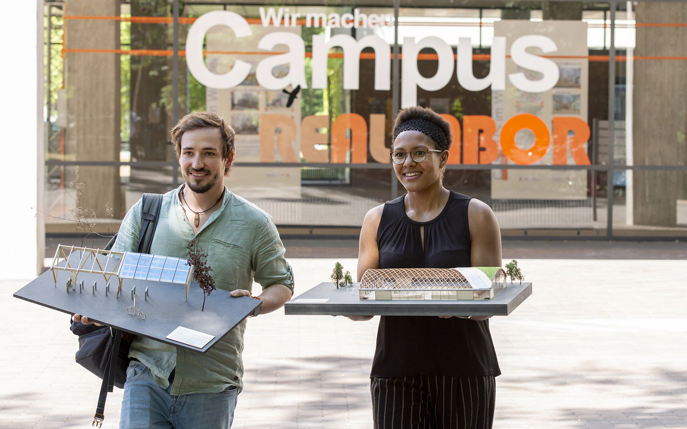 two students in front of the Civil Engineering building, carrying models of buildings