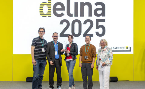 Das Bild zeigt fünf Personen, die auf einer Bühne vor einer großen Leinwand mit der Aufschrift „delina 2025“ stehen. In der Mitte hält Nadine Knobloch die Trophäe. Links von ihr stehen Dr. Oliver Langewitz und Prof. Dr. Matthias Wölfel, rechts Oskar Tanaka und Sünne Eichler. Alle lächeln in die Kamera, der Hintergrund ist hellgelb mit dem LEARNTEC-Logo unten rechts.