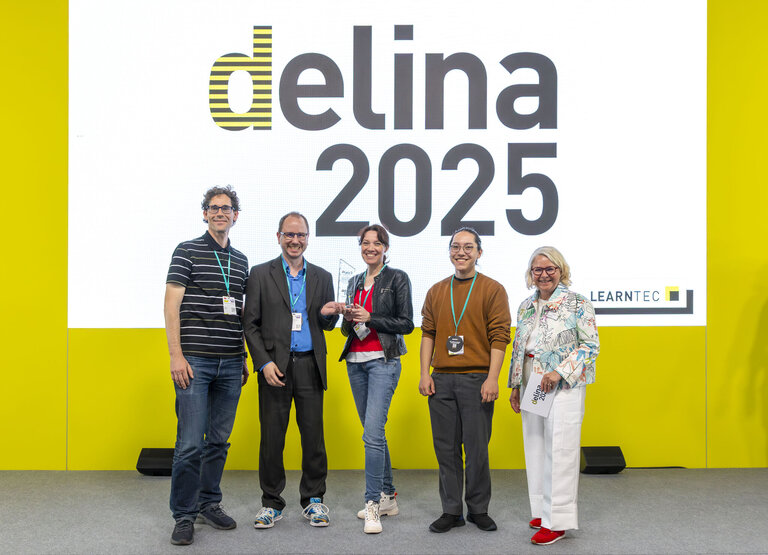Das Bild zeigt f&uuml;nf Personen, die auf einer B&uuml;hne vor einer gro&szlig;en Leinwand mit der Aufschrift &bdquo;delina 2025&ldquo; stehen. In der Mitte h&auml;lt Nadine Knobloch die Troph&auml;e. Links von ihr stehen Dr. Oliver Langewitz und Prof. Dr. Matthias W&ouml;lfel, rechts Oskar Tanaka und S&uuml;nne Eichler. Alle l&auml;cheln in die Kamera, der Hintergrund ist hellgelb mit dem LEARNTEC-Logo unten rechts.