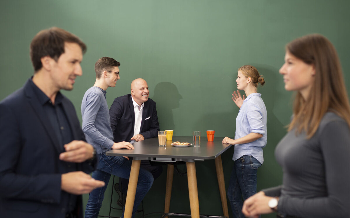 Zwei Menschen stehen vorne und drei hinten an einem Tisch und unterhalten sich. 