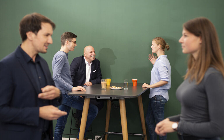 Zwei Menschen stehen vorne und drei hinten an einem Tisch und unterhalten sich. 