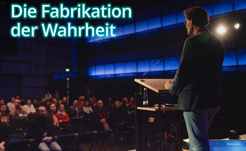 Prof. Dr. Matthias Wölfel stands at the lectern on the stage of the Media Theater at the ZKM Karlsruhe and speaks to an audience in a dark hall with blue lighting. Several people sit in rows in front of him and listen attentively. Dr. Wölfel is wearing a dark jacket and jeans. At the top left of the picture, in large, bright blue letters, is the text: "The Fabrication of Truth".