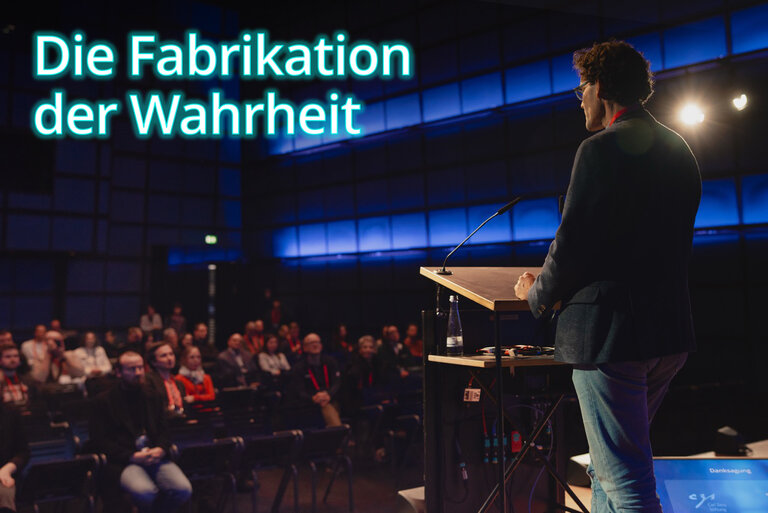 Prof. Dr. Matthias W&ouml;lfel steht am Rednerpult auf der B&uuml;hne des Medientheaters im ZKM Karlsruhe und spricht zu einem Publikum in einem dunklen Saal mit blauer Beleuchtung. Vor ihm sitzen mehrere Menschen in Reihen und h&ouml;ren aufmerksam zu. Dr. W&ouml;lfel tr&auml;gt ein dunkles Sakko und Jeans. Links oben im Bild steht in gro&szlig;en, leuchtend blauen Buchstaben der Text: &bdquo;Die Fabrikation der Wahrheit&ldquo;.