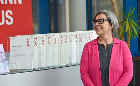 Eine Frau in pinkfarbenem Blazer steht lächelnd neben einer Reihe aufgestellter Glasurkunden mit der Aufschrift „QuantenPIONIER" und dem HKA-Logo.