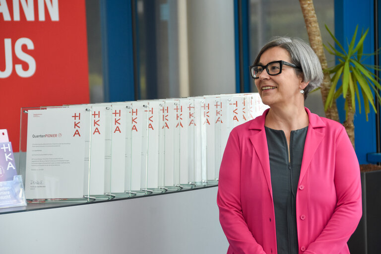 Eine Frau in pinkfarbenem Blazer steht lächelnd neben einer Reihe aufgestellter Glasurkunden mit der Aufschrift „QuantenPIONIER" und dem HKA-Logo.