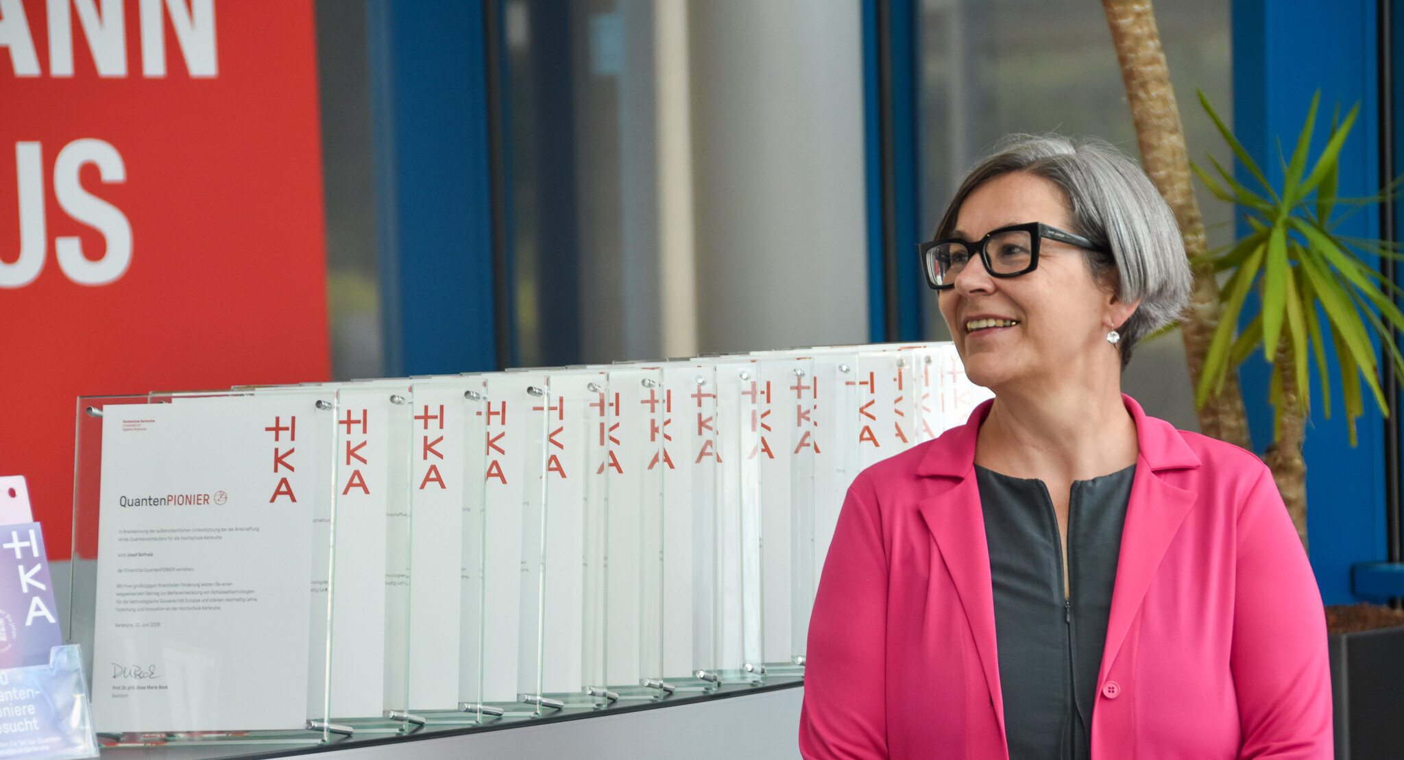Eine Frau in pinkfarbenem Blazer steht lächelnd neben einer Reihe aufgestellter Glasurkunden mit der Aufschrift „QuantenPIONIER" und dem HKA-Logo.