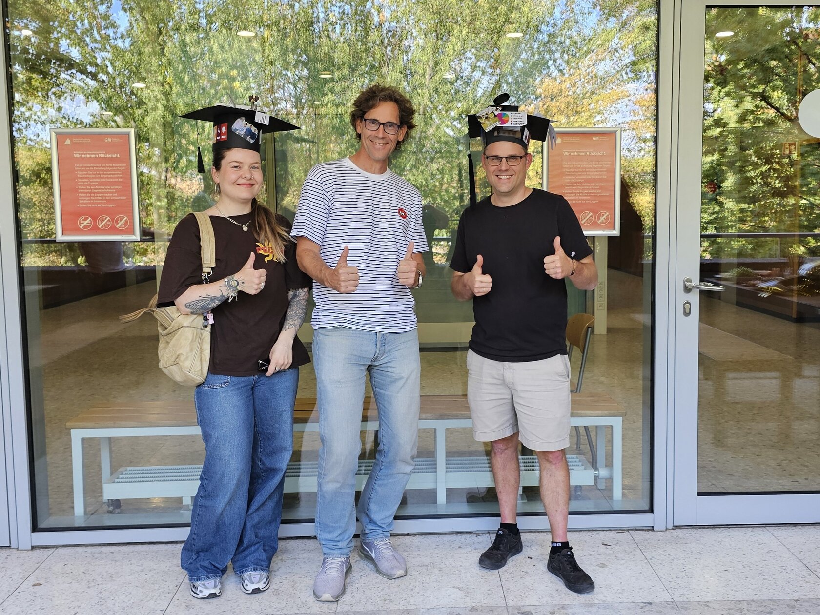 Auf dem Bild sieht man von links nach rechts, Melinda Braun, Matthias Wölfel und Thorsten Zyloswki. Melinda Braun und Thorsten Zylowski tragen beide ihre Doktorhüte. Alle drei zeigen ihre Daumen nach oben. Das Bild wurde vor einer Glasfront auf einem Balkon der HKA aufgenommen. 